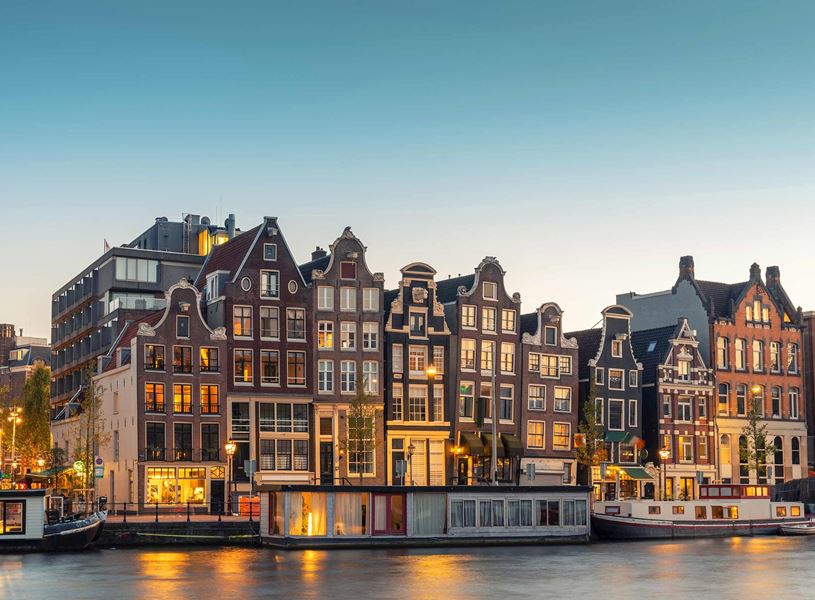 Gabled buildings along Amsterdam canal with moored houseboats illuminated by evening lights 