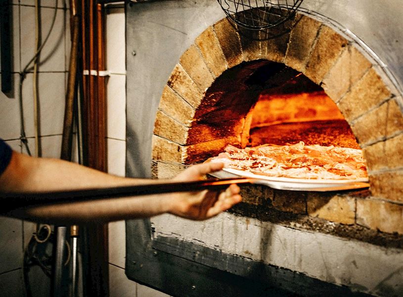Pizza being placed into traditional wood-fired brick oven with glowing embers 